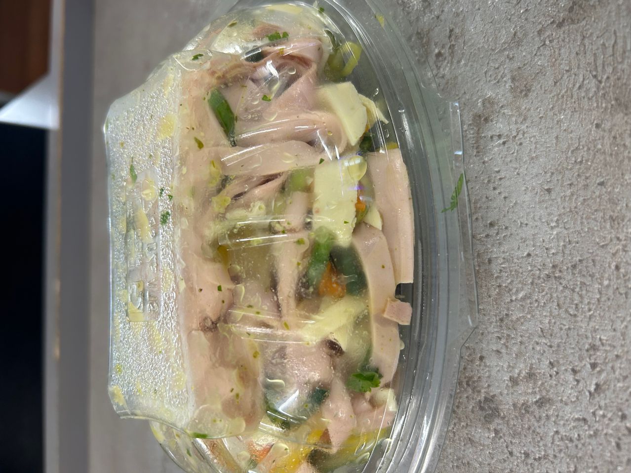 Ein Plastikbehälter enthält ein Gericht mit geschnittenem Fleisch, grünem Gemüse und gelbem Käse, abgedeckt mit durchsichtiger Plastikfolie.