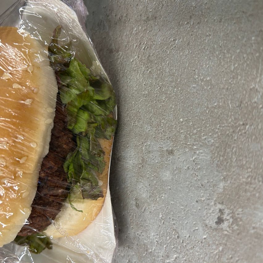 Ein in Plastik eingewickelter Hamburger mit Salat darauf, auf einem grauen strukturierten Hintergrund.