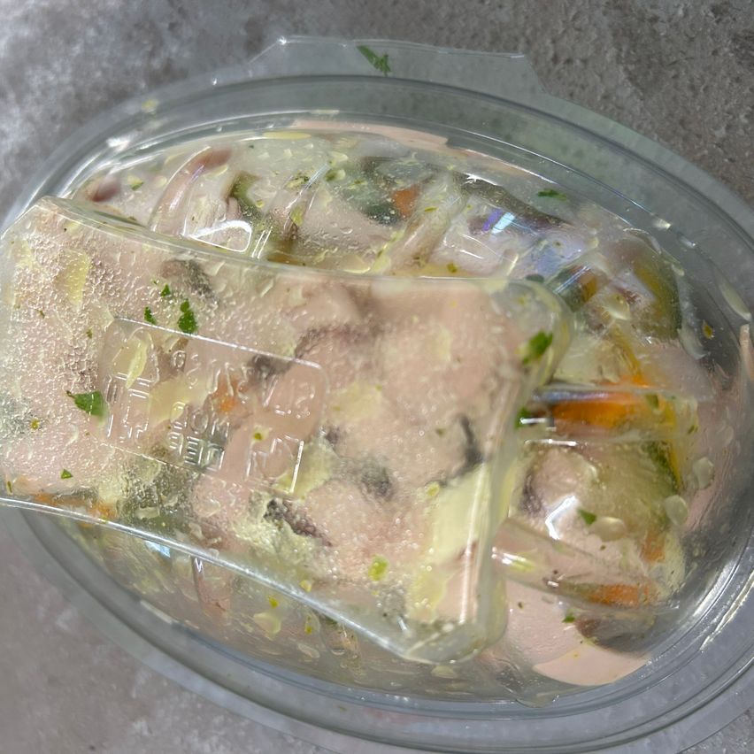 Ein durchsichtiger Plastikbehälter gefüllt mit einem Hühnchen-Gemüse-Salat, konserviert in einer Flüssigkeit. Das Gericht hat eine Mischung aus Hähnchen, Gemüse und Kräutern, die in klarer Flüssigkeit eingetaucht sind.