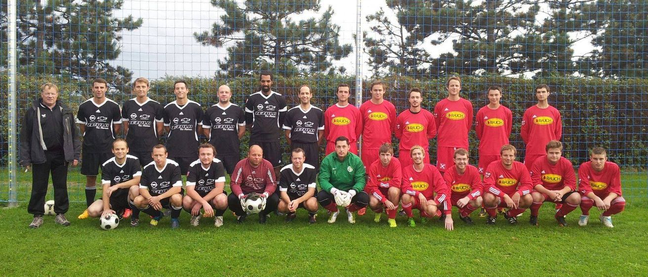 Eine Fußballmannschaft in roten und schwarzen Trikots posiert für ein Gruppenfoto vor einem Netzzaun, mit Bäumen im Hintergrund.
