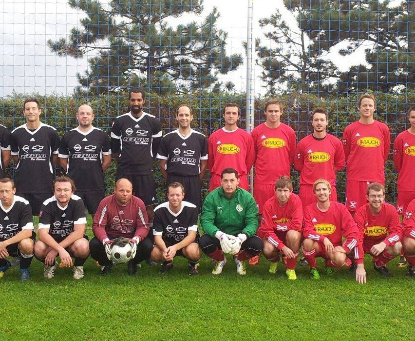 Eine Fußballmannschaft in roten und schwarzen Trikots posiert für ein Gruppenfoto vor einem Netzzaun, mit Bäumen im Hintergrund.