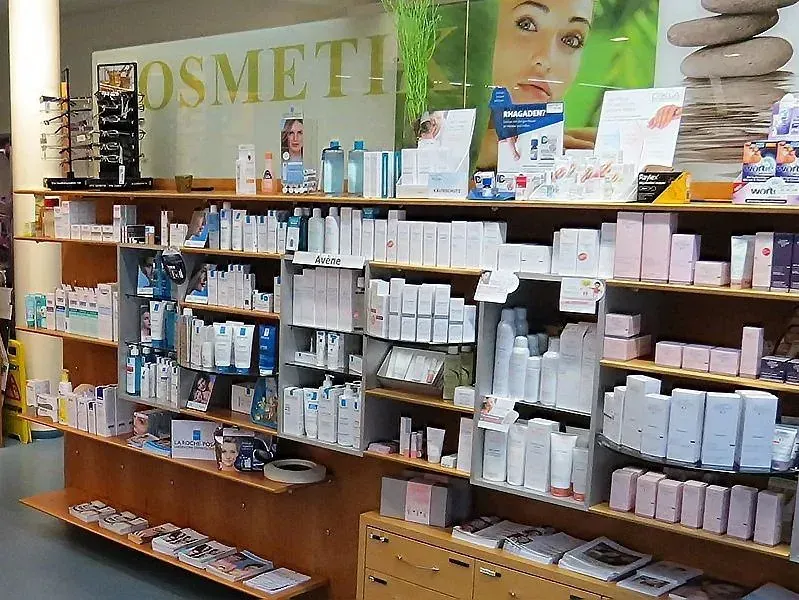 Ein Geschäft mit einem Regal voller verschiedener Schönheitsprodukte und einem Schild, das 'Kosmetik' steht. Es gibt auch einige Broschüren und Zeitschriften auf den unteren Regalen. Über dem Regal befindet sich ein Poster mit dem Gesicht einer Frau und dem Wort 'Kha'.