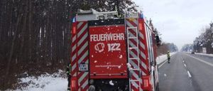 Ein Feuerwehrauto räumt Schnee von einem Baum am Rande einer verschneiten Straße, wobei eine Person in der Nähe und eine andere auf der Straße steht.