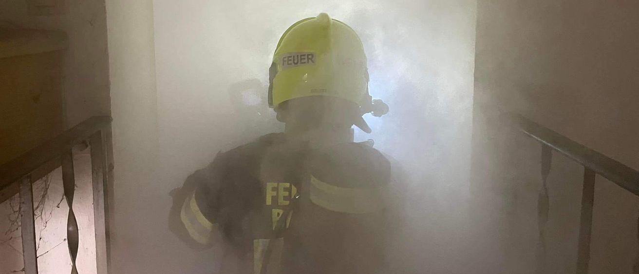 Ein Feuerwehrmann in einem gelben Helm mit dem Wort 'FEUER' steht in einem rauchgefüllten Raum, wahrscheinlich als Reaktion auf einen Brand.