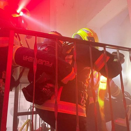 Drei Feuerwehrleute in Schutzausrüstung, mit Helmen, klettern eine Metallreling hinauf, wahrscheinlich als Reaktion auf einen Notfall. Einer hat eine Taschenlampe an.