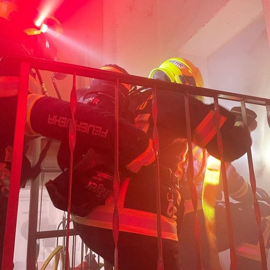 Drei Feuerwehrleute in Schutzausrüstung, mit Helmen, klettern eine Metallreling hinauf, wahrscheinlich als Reaktion auf einen Notfall. Einer hat eine Taschenlampe an.