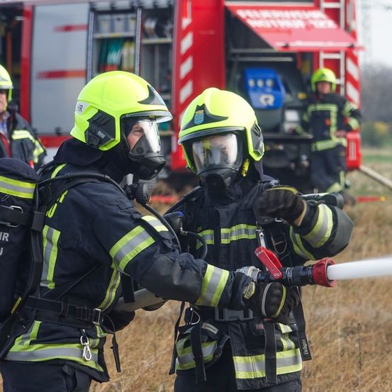 Feuerwehrleute in leuchtend gelben Helmen und schwarzen Uniformen üben mit einem Schlauch, vor einem roten Feuerwehrauto stehend.