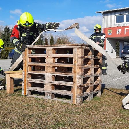 Zwei Feuerwehrleute trainieren auf einer Übungsplattform und ziehen einen Schlauch über einen Stapel Holzpaletten. Im Hintergrund befindet sich ein Gebäude mit einem Schild, auf dem 'F' steht.