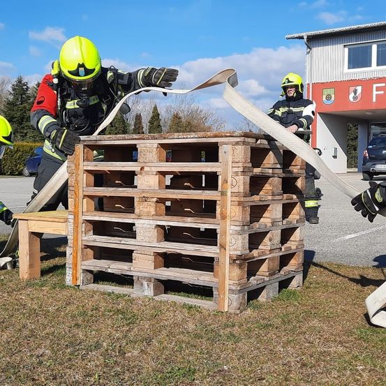Zwei Feuerwehrleute trainieren auf einer Übungsplattform und ziehen einen Schlauch über einen Stapel Holzpaletten. Im Hintergrund befindet sich ein Gebäude mit einem Schild, auf dem 'F' steht.