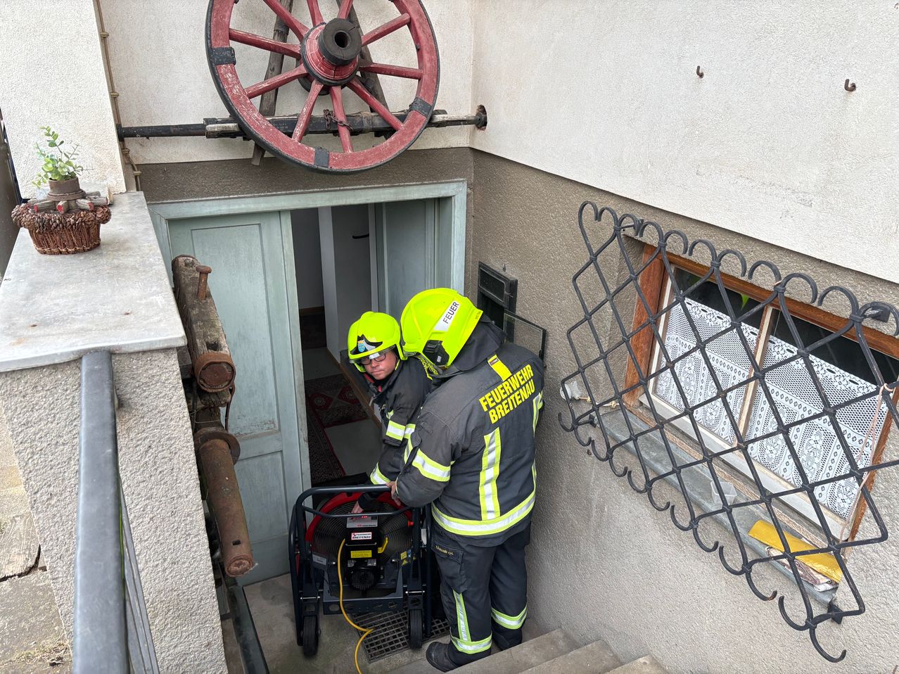 Zwei Feuerwehrleute mit gelben Helmen sind auf den Treppen, einer schiebt einen fahrbaren Generator und der andere hält einen Schlauch in der Nähe einer offenen Tür. Ein altes rotes Rad ist über der Tür montiert.