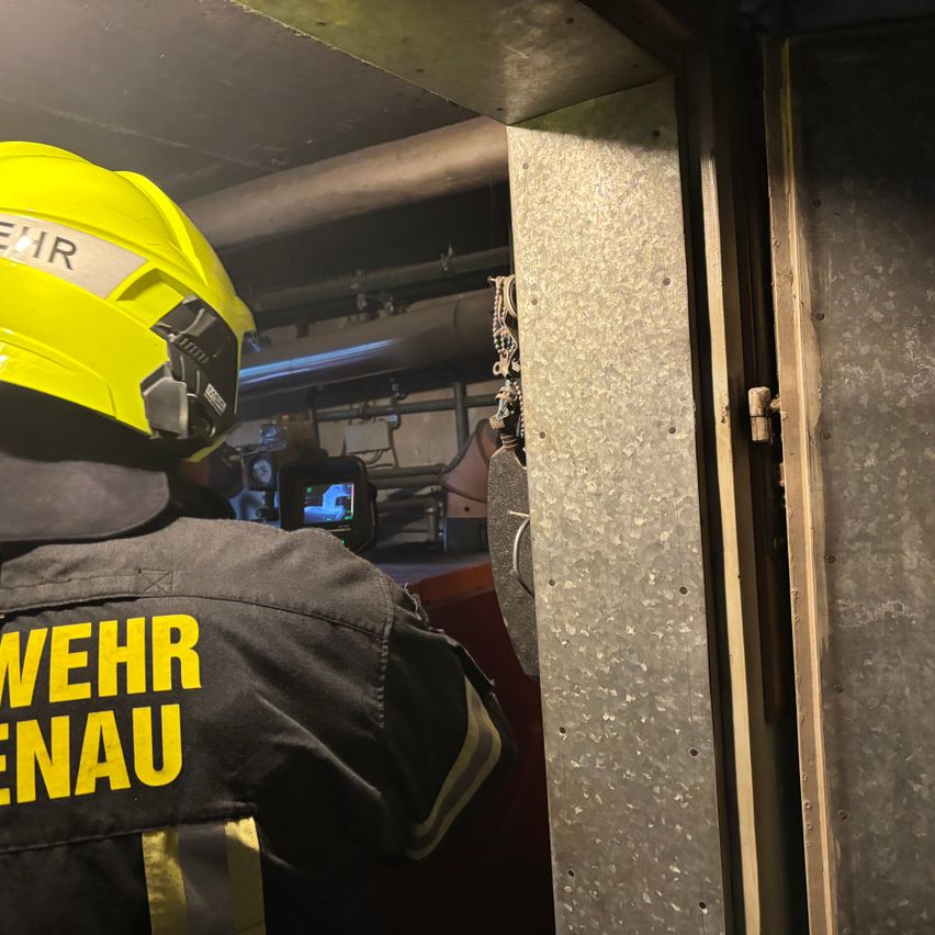Ein Feuerwehrmann mit einem gelben Helm, auf dem 'WEHR' steht, steht in einem Raum mit Metallwänden, dessen Tür offen ist und der möglicherweise inspiziert wird.