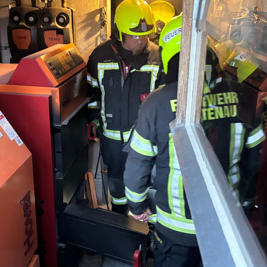 Feuerwehrleute stehen in einem Raum mit Industriegeräten, tragen gelbe Helme und Schutzanzüge. Die Geräte haben rote und orangefarbene Farben, mit dem Wort Hoval darauf.