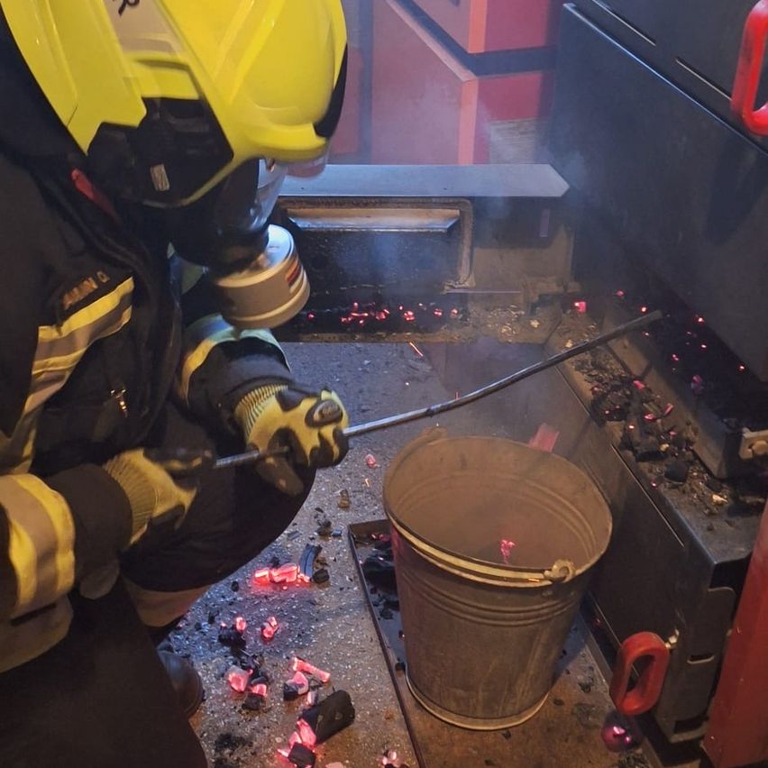 Ein Feuerwehrmann kniet mit einer Schaufel neben einem Feuer und hält einen Eimer mit rotem Griff. Die Szene ist gefüllt mit glühenden Kohlen und Rauch.