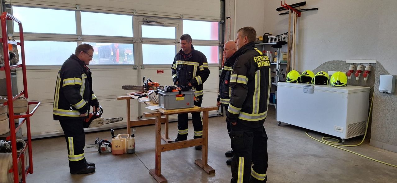 Three firefighters stand in a room, discussing near a table with a chainsaw and toolbox. The room has a garage door, windows, and storage shelves.