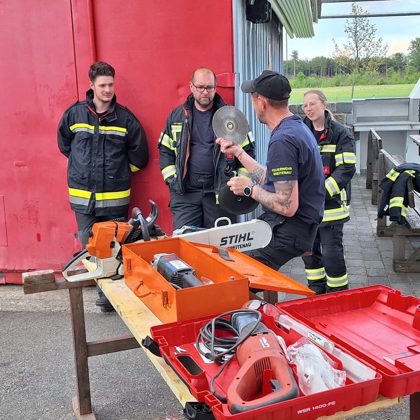 Firefighters in uniforms are gathered around a table with orange tool boxes and chainsaws. A man holds a disc. They stand next to a red building with a grassy field in the background.