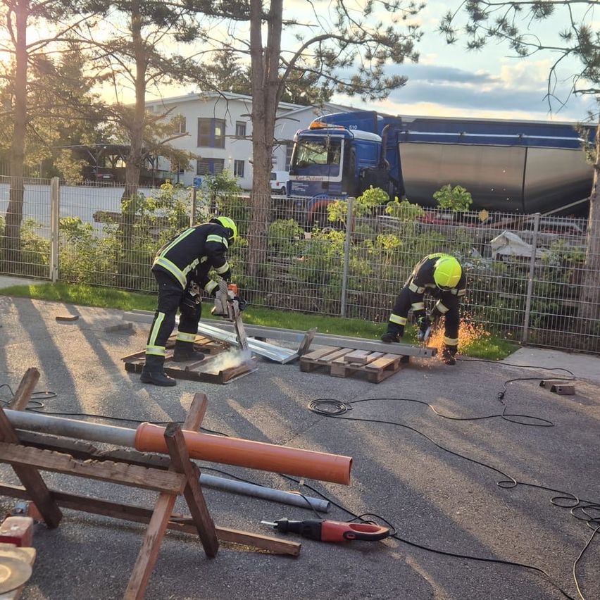 Two firefighters are cutting metal on the ground, with a blue tanker truck parked behind a fence. The area is surrounded by trees, and a building is visible in the background.