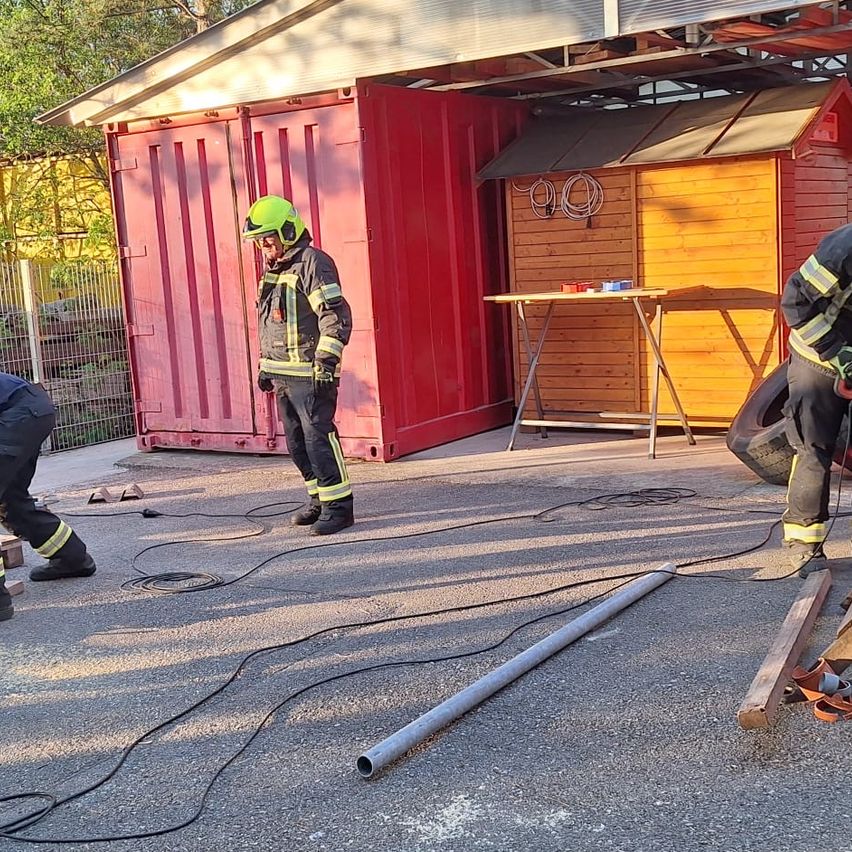 Three firefighters are outdoors with red containers and a wooden shed. One is standing, another bent over, and the third by the shed. Wires and tools are scattered.