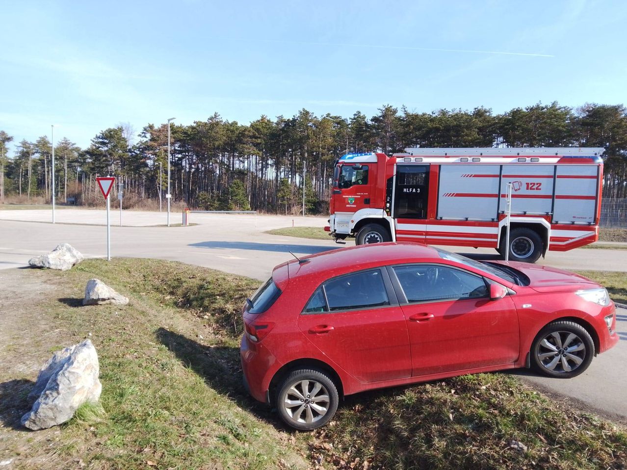 Ein rotes Auto ist am Straßenrand geparkt, mit einem Feuerwehrwagen dahinter. Ein dreieckiges Schild und ein Pfosten befinden sich auf der linken Seite.