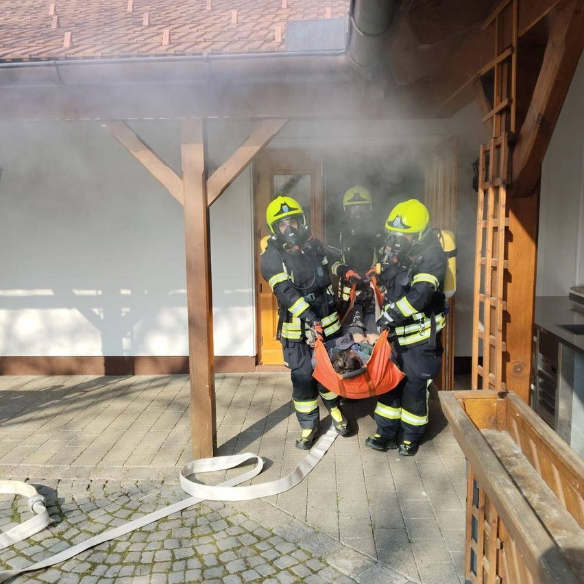 Drei Feuerwehrleute tragen eine Person auf einer Trage aus einem rauchgefüllten Raum. Der Raum befindet sich draußen mit einer Holzstruktur und einem Ziegelboden.