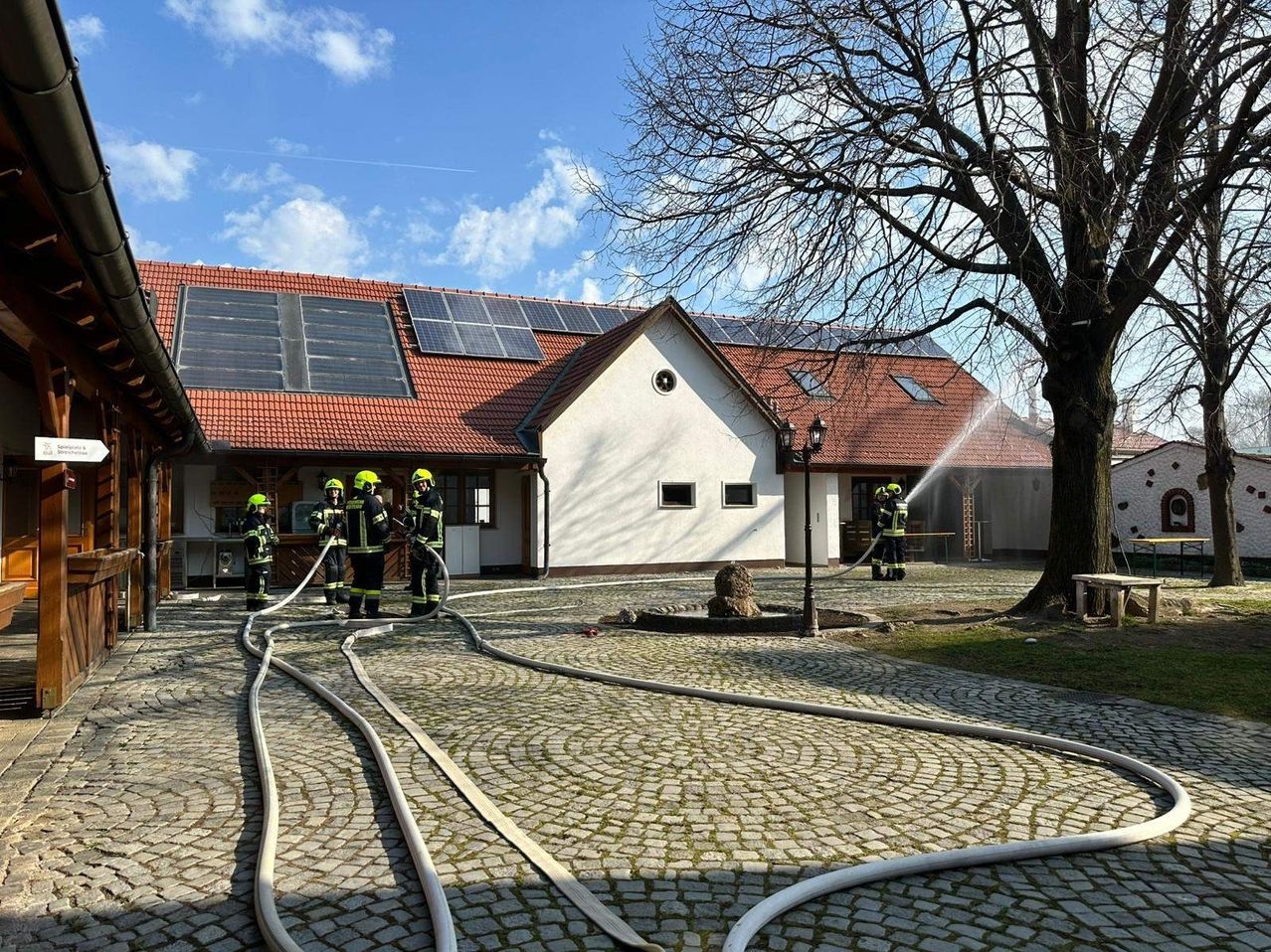 Feuerwehrleute sprühen Wasser auf ein Gebäude mit Solarpanelen auf dem Dach. Das Gebäude hat ein rotes Dach und ist von einem Kopfsteinpflaster umgeben.