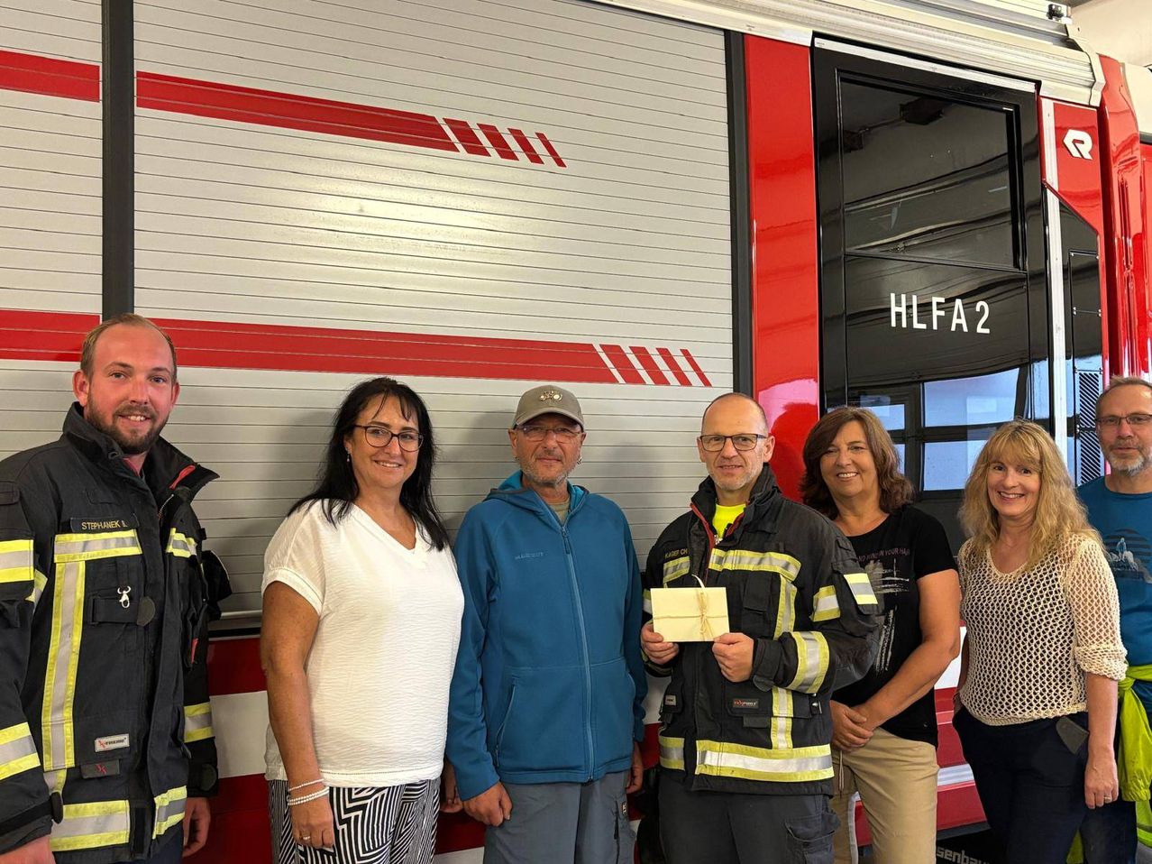 Eine Gruppe von Menschen steht vor einem Feuerwehrauto, ein Mann hält ein Geschenk. Das Feuerwehrauto hat rote Streifen und die Aufschrift 'HLFA2' an der Tür.