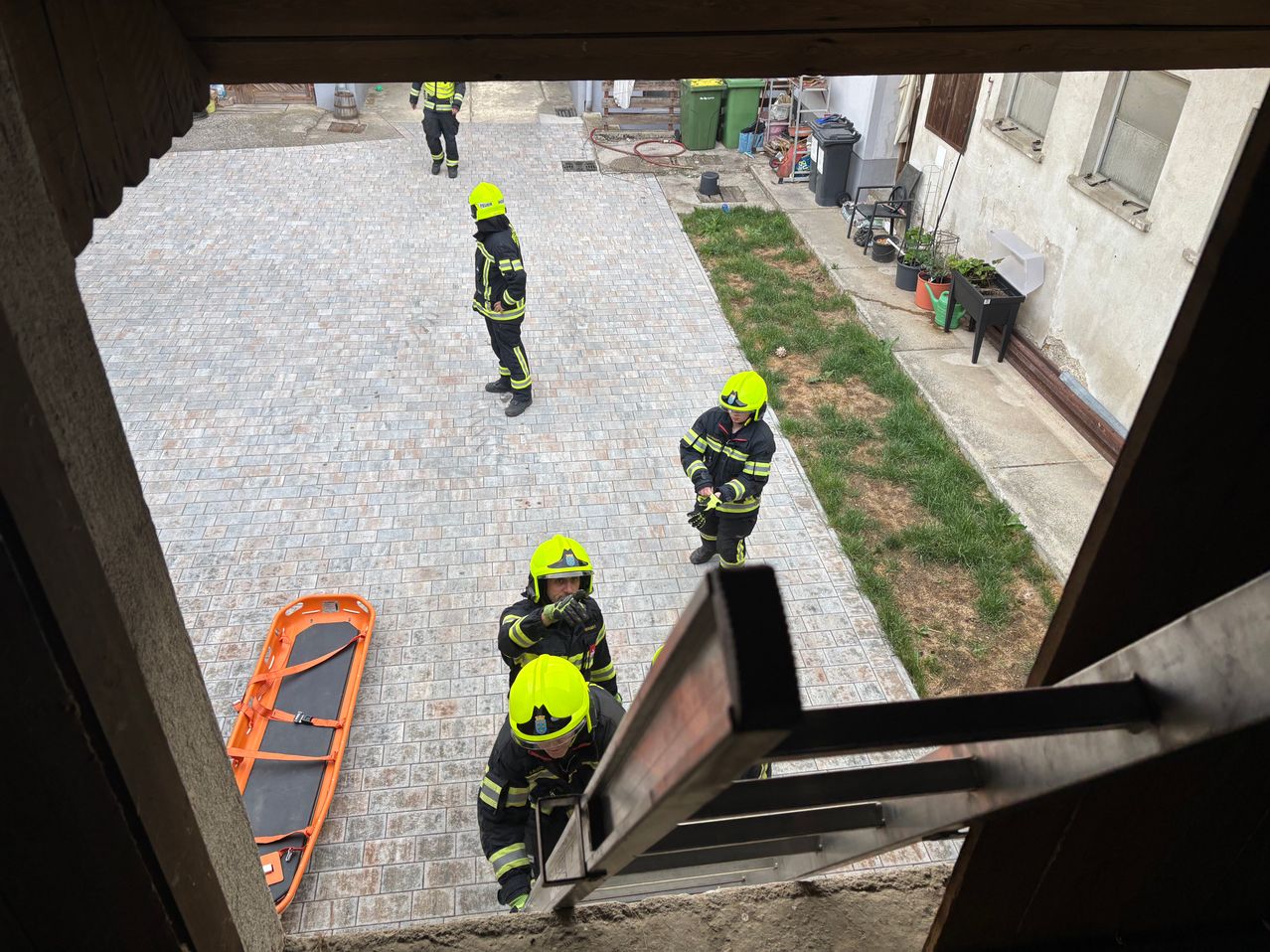 Feuerwehrleute versammeln sich auf einem Hof, einer klettert eine Leiter hinunter, zwei andere stehen und einer geht weg. Eine Trage liegt auf dem Boden.