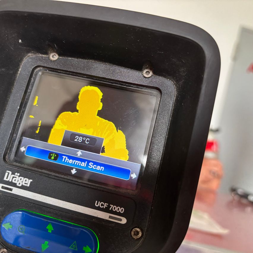 Nahaufnahme eines Thermoscan-Geräts, das eine gelbe Silhouette einer Person mit einer Temperaturanzeige von 28°C zeigt. Das Gerät ist mit 'Drager' und Modellnummer UCF 7000 beschriftet.