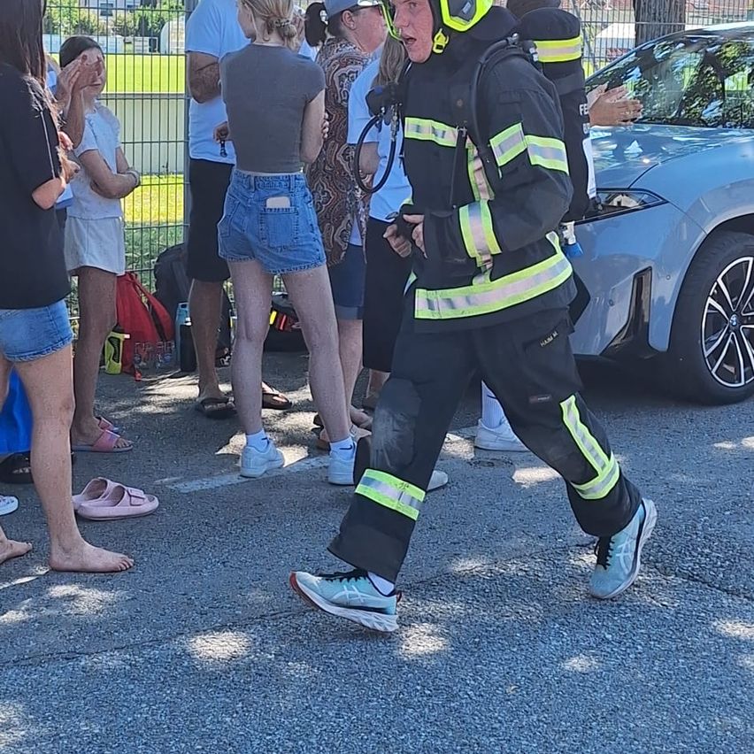 Ein Feuerwehrmann in Uniform geht an einer Gruppe von Menschen vorbei, ein Auto ist in der Nähe geparkt.