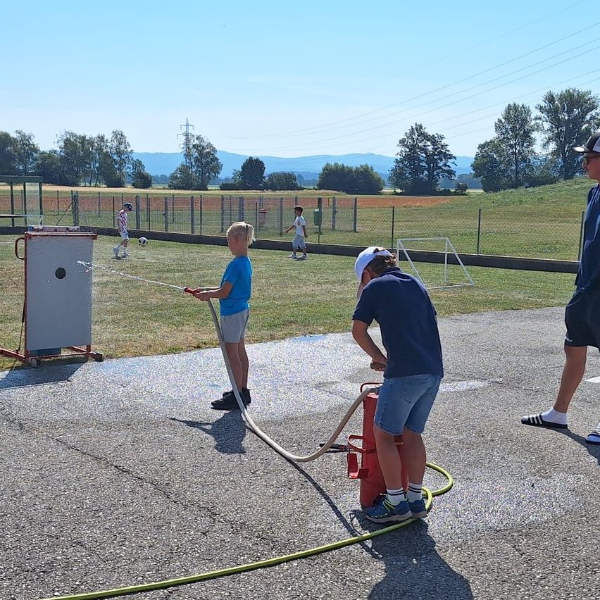 Zwei Kinder reinigen den Boden mit einem Wasserschlauch. Ein Kind hält den Schlauch und das andere steht auf einem roten Eimer. Sie sind auf einem Betonboden. Es gibt andere Kinder, die Fußball spielen.