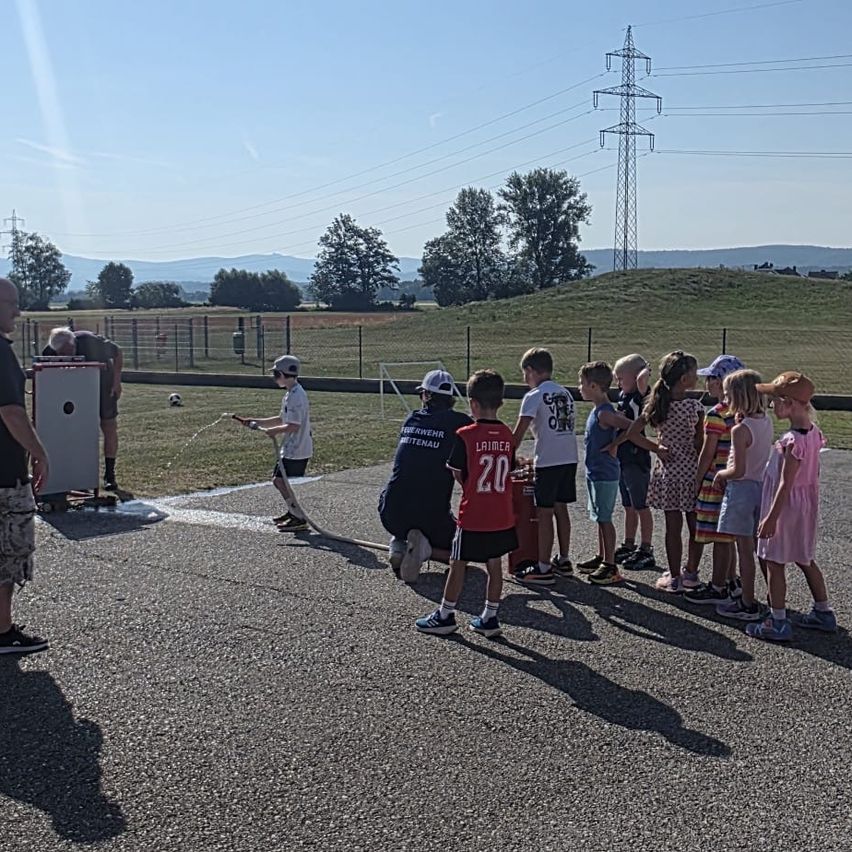 Eine Gruppe von Kindern und Erwachsenen versammelt sich auf einer gepflasterten Fläche, mit einem Fußball im Hintergrund. Ein Mann sprüht Wasser.
