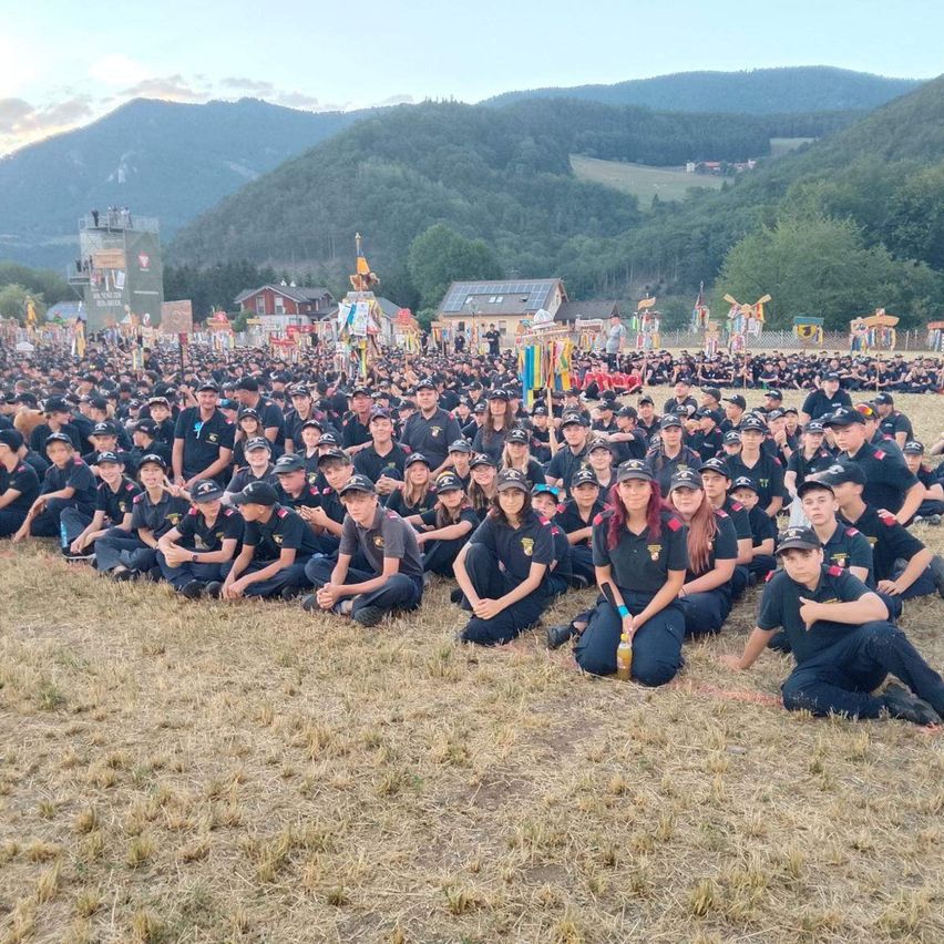 Eine große Gruppe von Menschen in Uniform sitzt auf dem Boden in einem Feld, mit Bergen und einem klaren Himmel im Hintergrund.
