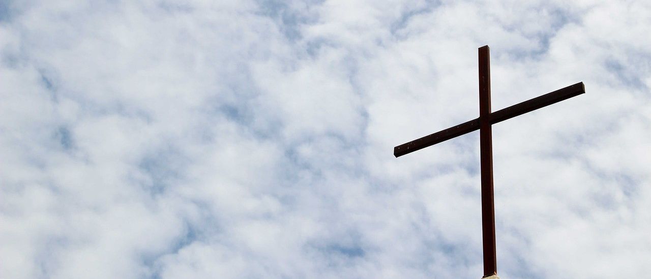 Ein großes Kreuz steht auf einem Gebäude, mit einem bewölkten blauen Himmel im Hintergrund. Das Kreuz ist aus Metall und hat eine braune Oberfläche.