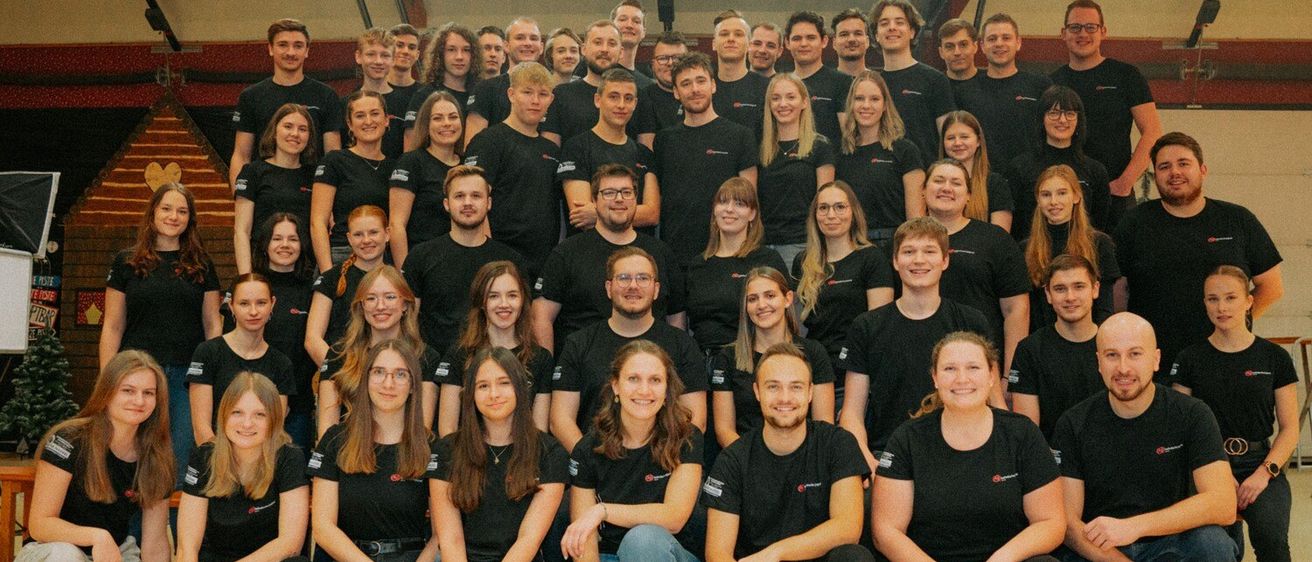 Eine Gruppe von Menschen in passenden schwarzen T-Shirts mit Logos posiert für ein Foto. Sie sitzen und stehen auf einem Holzboden.
