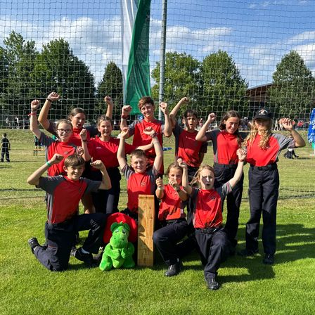 Eine Gruppe von Kindern und eine grüne Stofftier posieren für ein Foto auf einem Grasfeld mit einem Netz im Hintergrund.