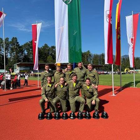 Eine Gruppe von Militärangehörigen in grünen Uniformen posiert auf einer roten Laufbahn für ein Foto mit Helmen vor sich. Dahinter stehen Fahnen auf Stangen.