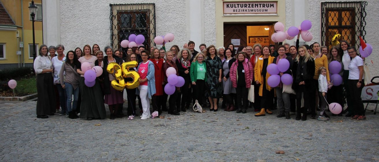 Eine Gruppe von Menschen steht vor einem Gebäude mit der Aufschrift 'Kulturzentrum Bezirkmuseum'. Sie halten alle Luftballons und tragen Freizeitkleidung. Einige von ihnen tragen eine Brille und halten einen goldenen Ballon mit der Aufschrift '35'.
