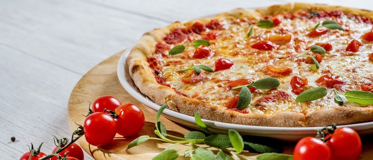 Eine ganze Pizza mit geschmolzenem Käse, Tomatenscheiben und Salbeiblättern auf einem Holztablett mit Kirschtomaten und Pfefferkörnern auf einem Holztisch.