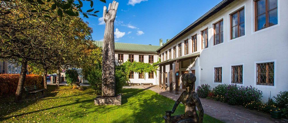 Eine Skulptur einer Frau sitzt auf einer Bank in einem Innenhof mit einer großen hölzernen Handplastik und einem Gebäude mit vielen Fenstern.