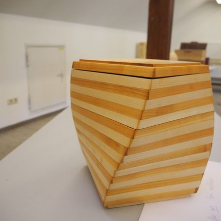 Eine Holzkiste mit mehreren Schubladen steht auf einem weißen Tisch. Die Kiste ist aus Holz und hat ein geometrisches Design. Sie befindet sich in einem Raum mit weißer Wand und Tür.