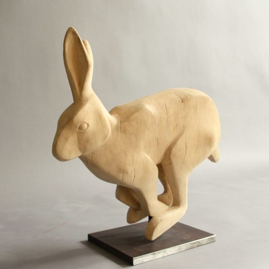 Eine hölzerne Skulptur eines Kaninchens in springender Haltung, mit detaillierten Ohren und Pfoten, auf einer dunklen Basis ausgestellt.