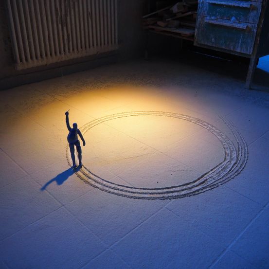 Ein Mann steht in einem Kreis in einem Raum mit gelbem Licht. Schatten und Spuren sind auf dem Boden sichtbar. Ein Heizkörper und ein Schrank befinden sich im Hintergrund.