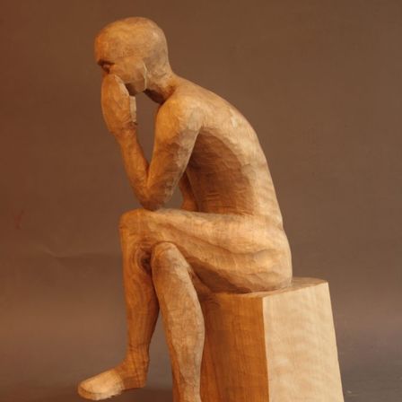 Eine Holzskulptur einer Person, die mit nachdenklichem Gesichtsausdruck auf einer Kiste sitzt. Die Figur ist aus einem Stück Holz geschnitzt.
