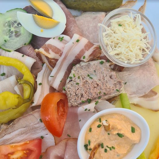 Eine Auswahl an geschnittenem Fleisch, Eiern, Gurken und Paprika ist auf einem Teller angerichtet. Schüsseln mit geriebenem Käse und Dip begleiten das Angebot.