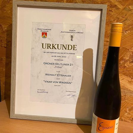 Ein gerahmtes Zertifikat, das neben einer Flasche Wein auf einer Holzoberfläche ausgestellt ist. Das Zertifikat trägt die Aufschrift 'Urkunde' und ist vom Bürgermeister unterzeichnet.