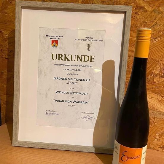 Ein gerahmtes Zertifikat, das neben einer Flasche Wein auf einer Holzoberfläche ausgestellt ist. Das Zertifikat trägt die Aufschrift 'Urkunde' und ist vom Bürgermeister unterzeichnet.