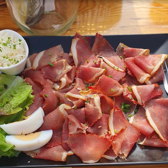 Ein schwarzer Teller mit Prosciutto-Schinken, Salat und gekochten Eiern. Ein Glas mit Wasser und ein Glas mit einem Löffel auf dem Tisch.