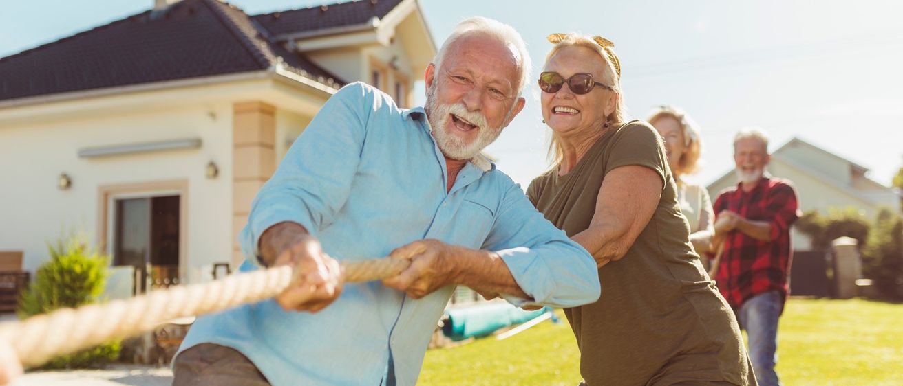 Ein älteres Paar streitet sich spielerisch beim Tauziehen an einem sonnigen Tag vor einem Haus.