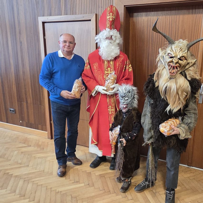 Ein Mann in einem blauen Pullover steht neben einer Person in Santa Claus-Kostüm und einer Person in einem hörnertragenden Dämonenkostüm. Sie halten Brot.