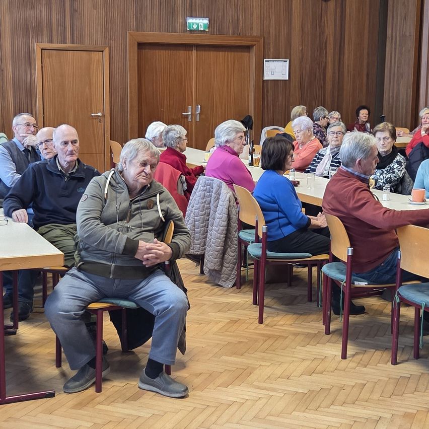 Eine Gruppe älterer Menschen sitzt in einem Raum mit Holzwänden und -böden, möglicherweise bei einer Versammlung. Einige sitzen, andere stehen, und es gibt Tische mit Gegenständen darauf.