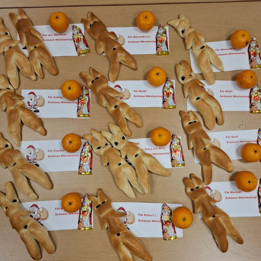 Eine Sammlung dekorativer Lebkuchenmänner mit verschiedenen Namen wie Petra, Claudia und Anja, jeweils begleitet von einer Orange und einer Schokoladenfigur des Weihnachtsmanns, auf einer Holzoberfläche angeordnet.